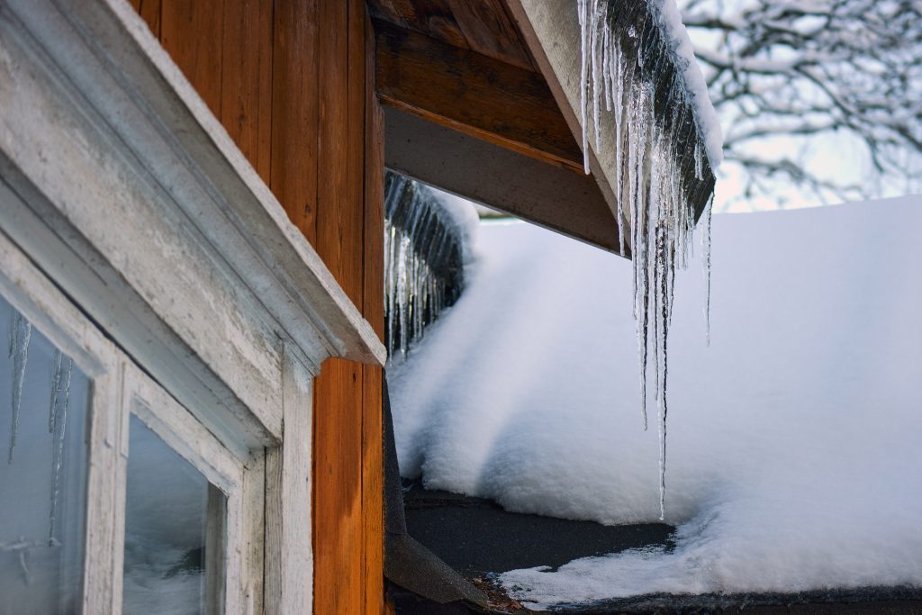 A close-up view of thick icicles and heavy snow accumulation on a wooden roof eave, illustrating what causes an ice dam on a roof during a Western New York winter.